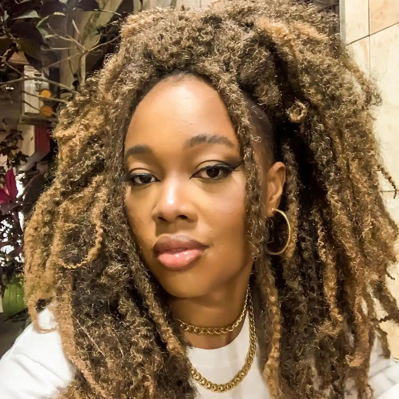 A woman with Marley hair styled in highlighted dreadlocks, wearing gold hoop earrings, a white top, and layered gold necklaces, gazing at the camera with a soft smile.