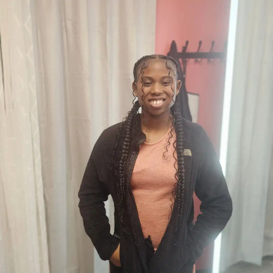 Person wearing a black cardigan over a pink shirt with braided hair, standing in front of a curtain.