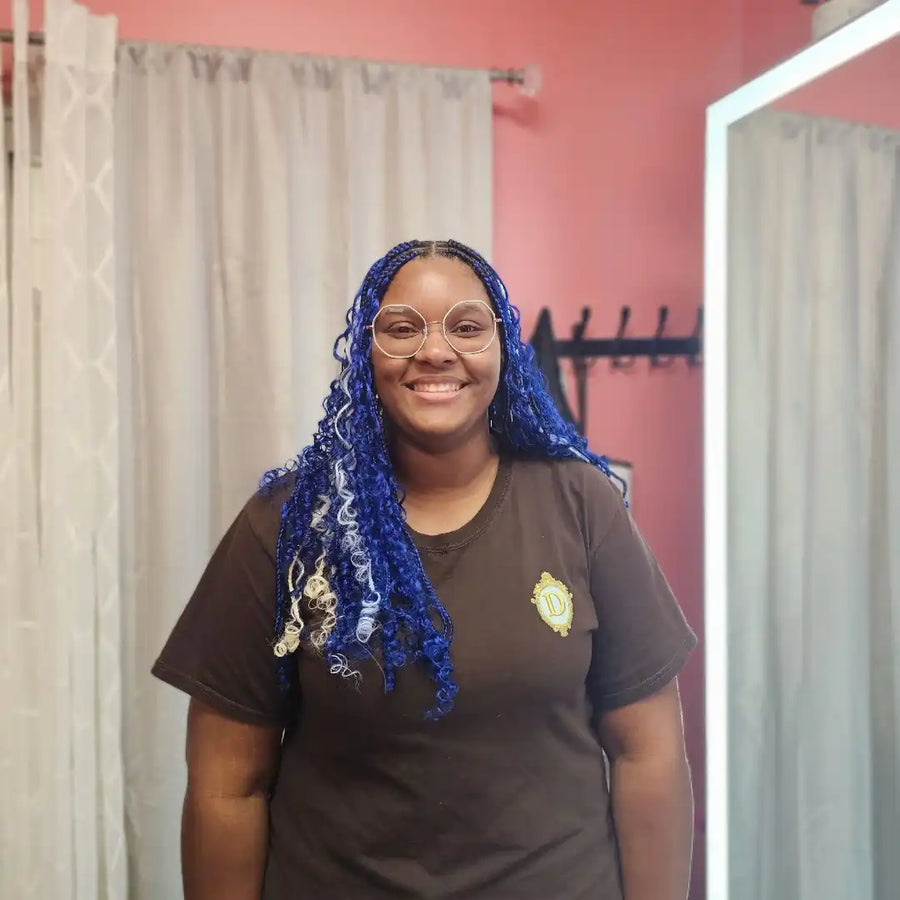 Person with blue hair wearing a brown shirt with a logo in a room with pink walls and curtains.