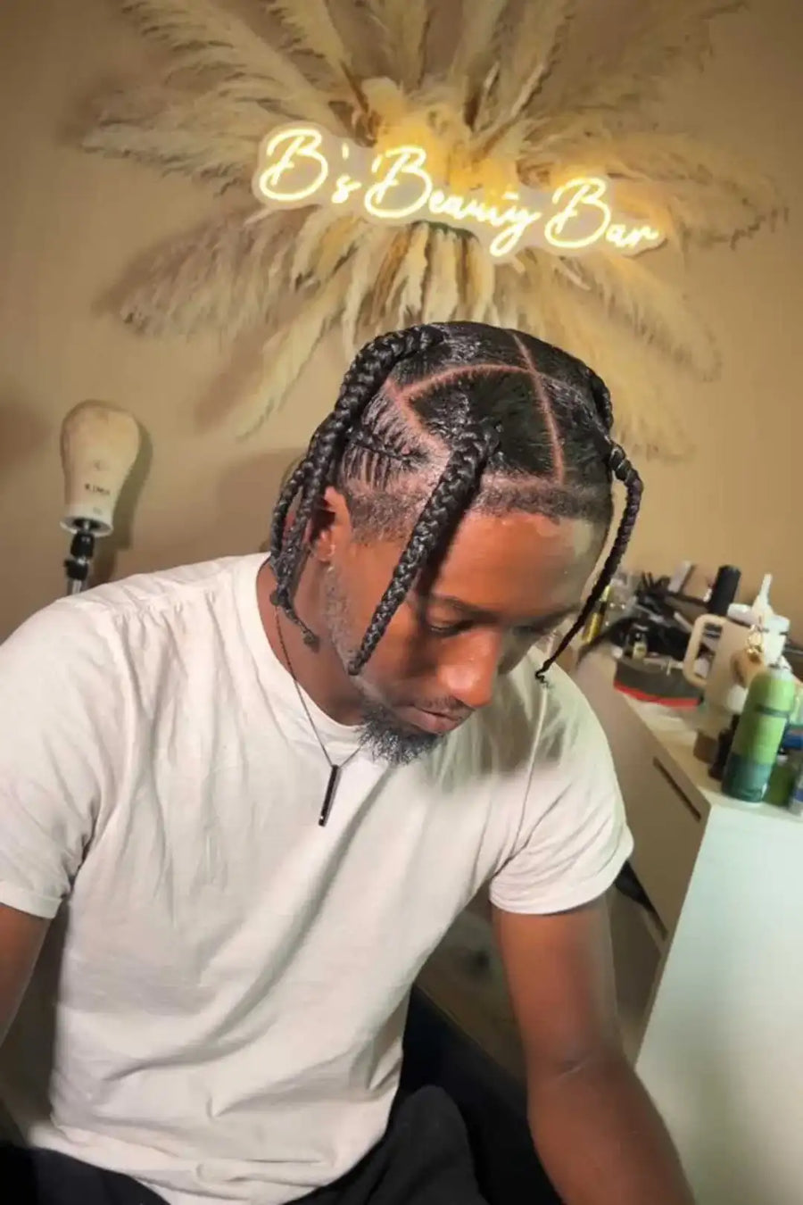 Man with styled hair in a salon setting with 'B's Braiding Bar' sign.