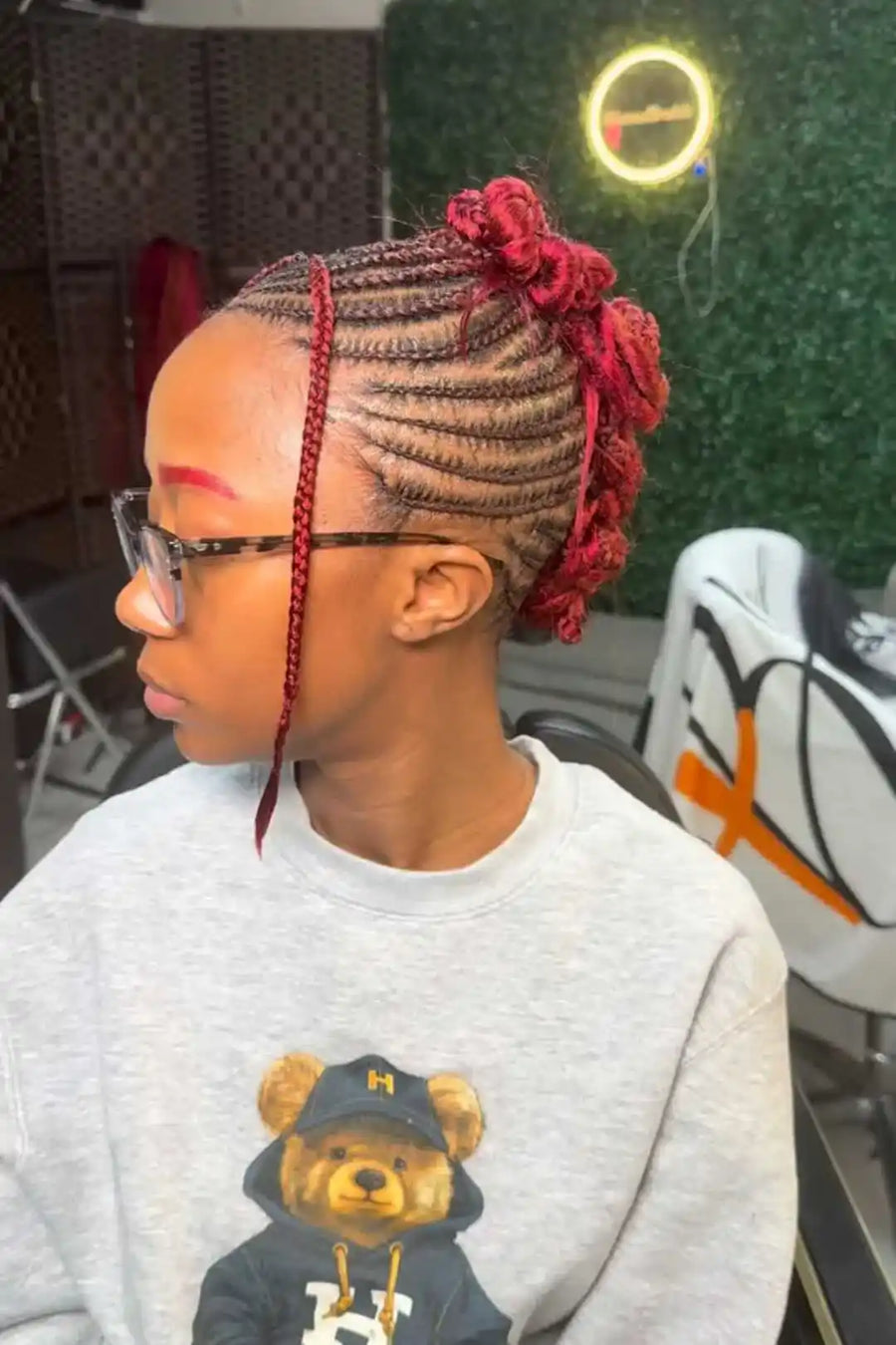 Person with braided hair styled with red accents, wearing a gray sweatshirt with a bear graphic.