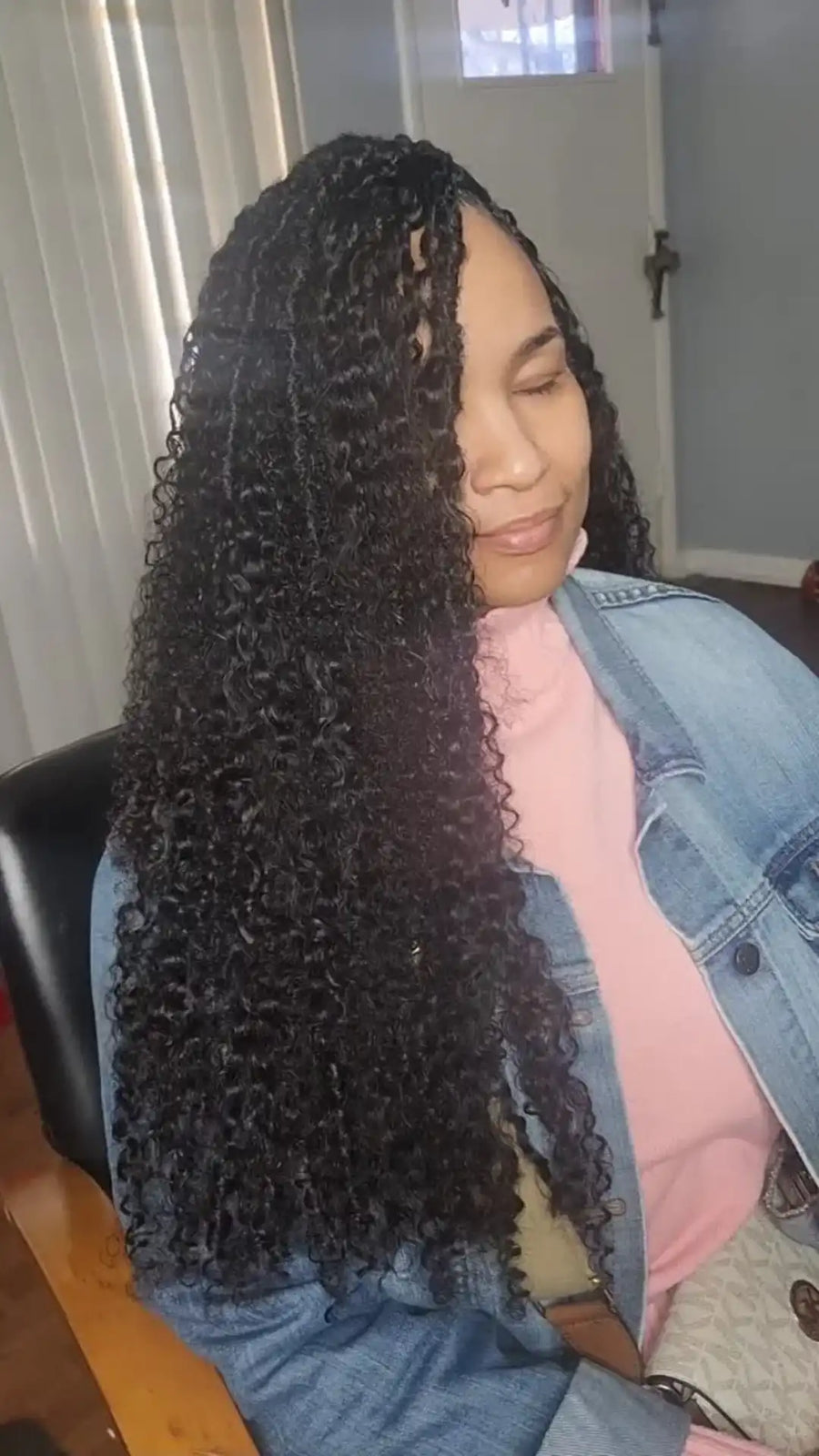 Person with long, curly hair sitting indoors wearing a denim jacket and pink shirt.
