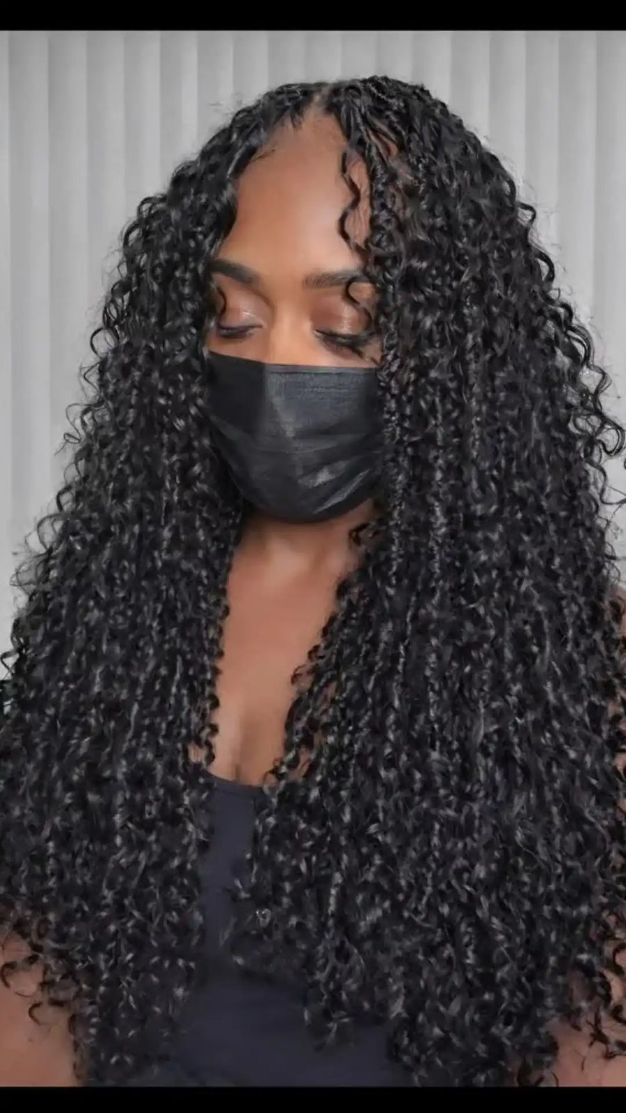 Person with long, curly black hair wearing a black face mask against a neutral background