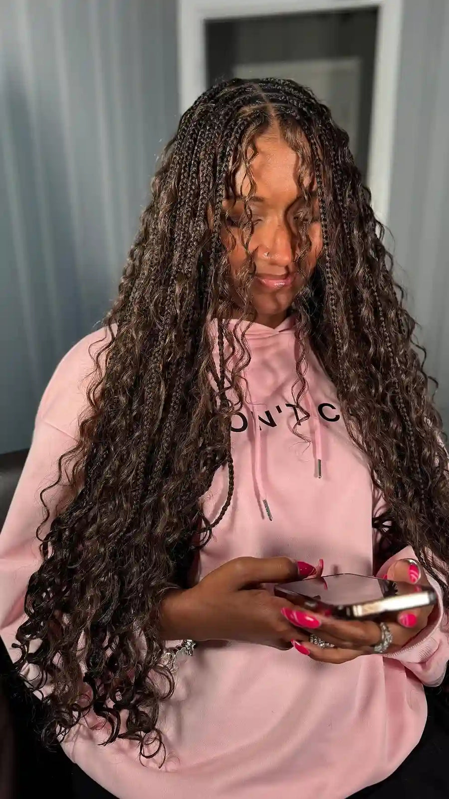 Person with long braided hair wearing a pink hoodie, holding a phone.
