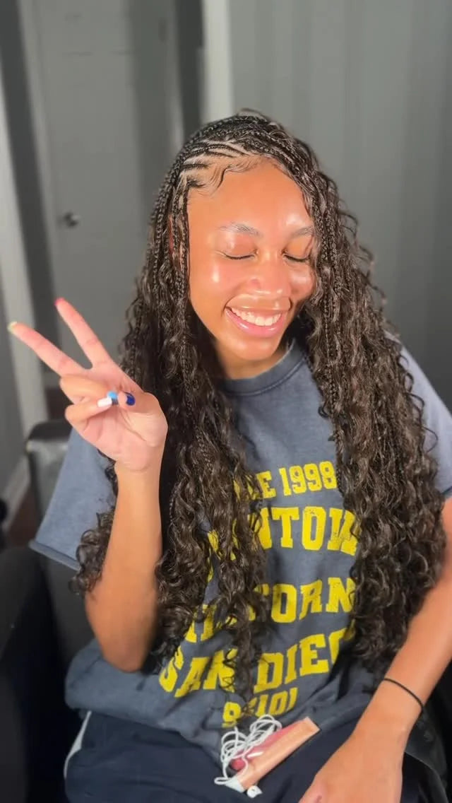 Person with long braided hair wearing a blue t-shirt with yellow text, making a peace sign.