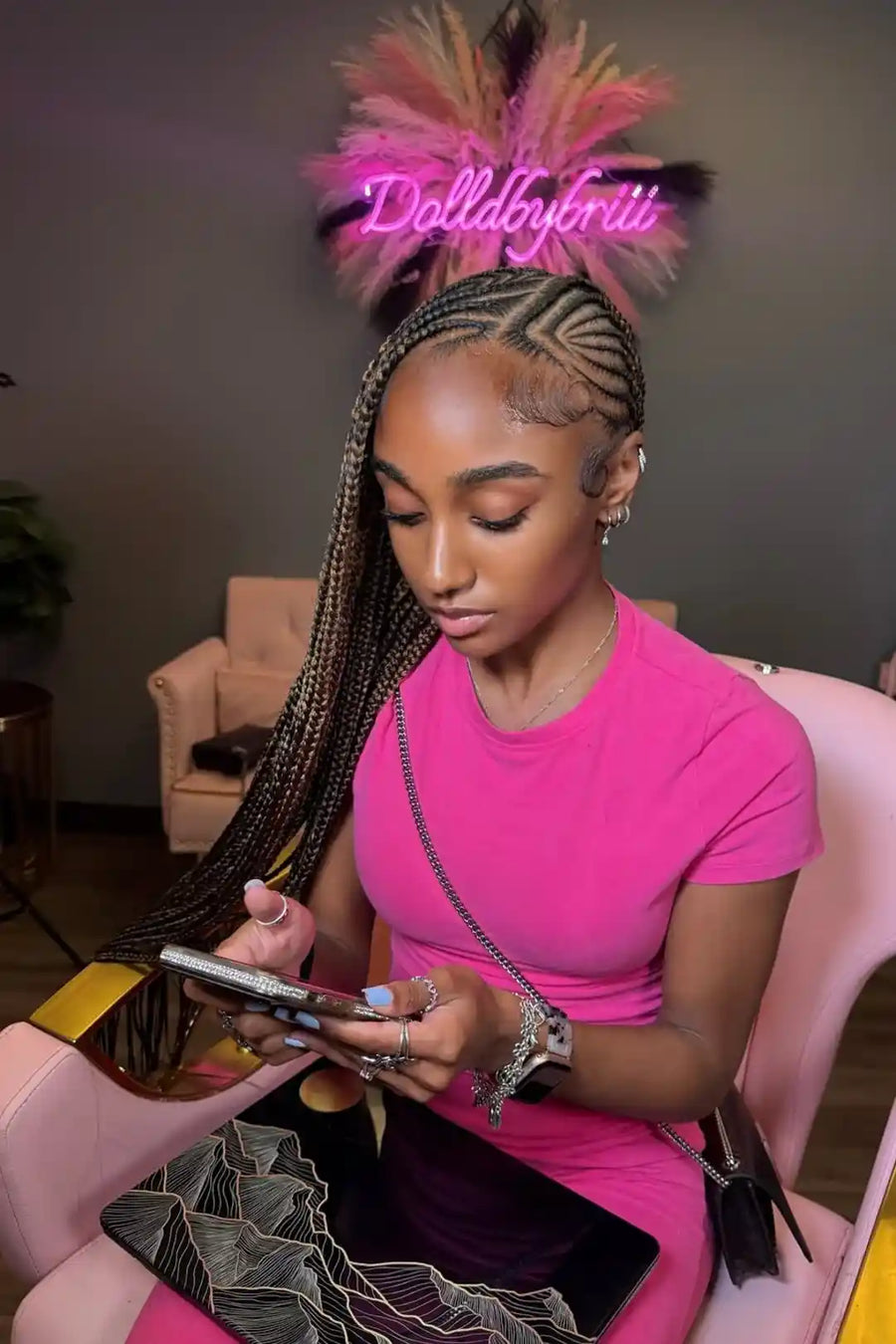 Woman in a pink dress sitting on a pink chair, using a phone with 'Dollabybri' branding in the background.