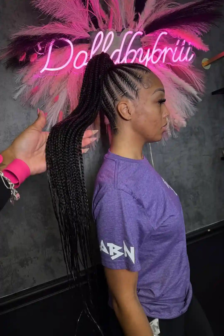 Person with braided hair and pink feathered headpiece in front of a neon sign.
