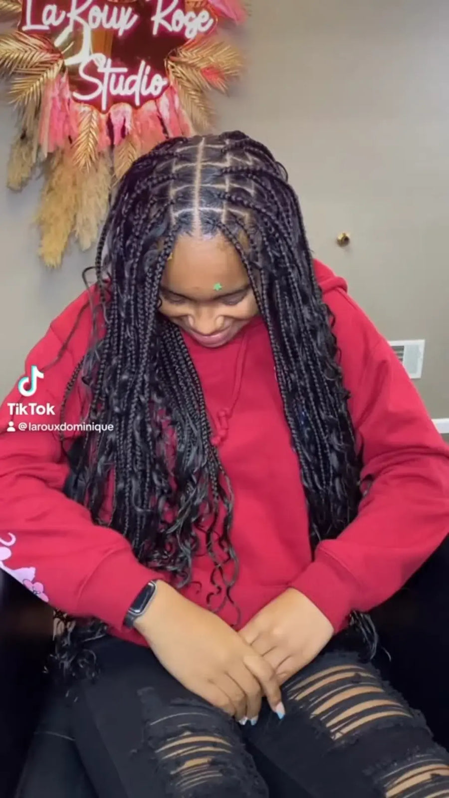 A woman with long black box braids sits indoors wearing a bright pink hoodie and ripped black jeans. The background shows a wall with a decorative wreath and a sign reading 'La Kouy Kose Studio', plus a TikTok watermark on the left.