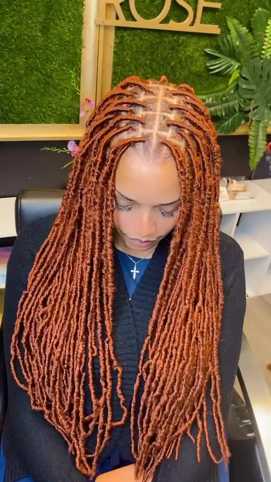 A person wearing long, coppery orange dreadlocks or box braids, seated indoors with a green wall and decorative plants in the background.