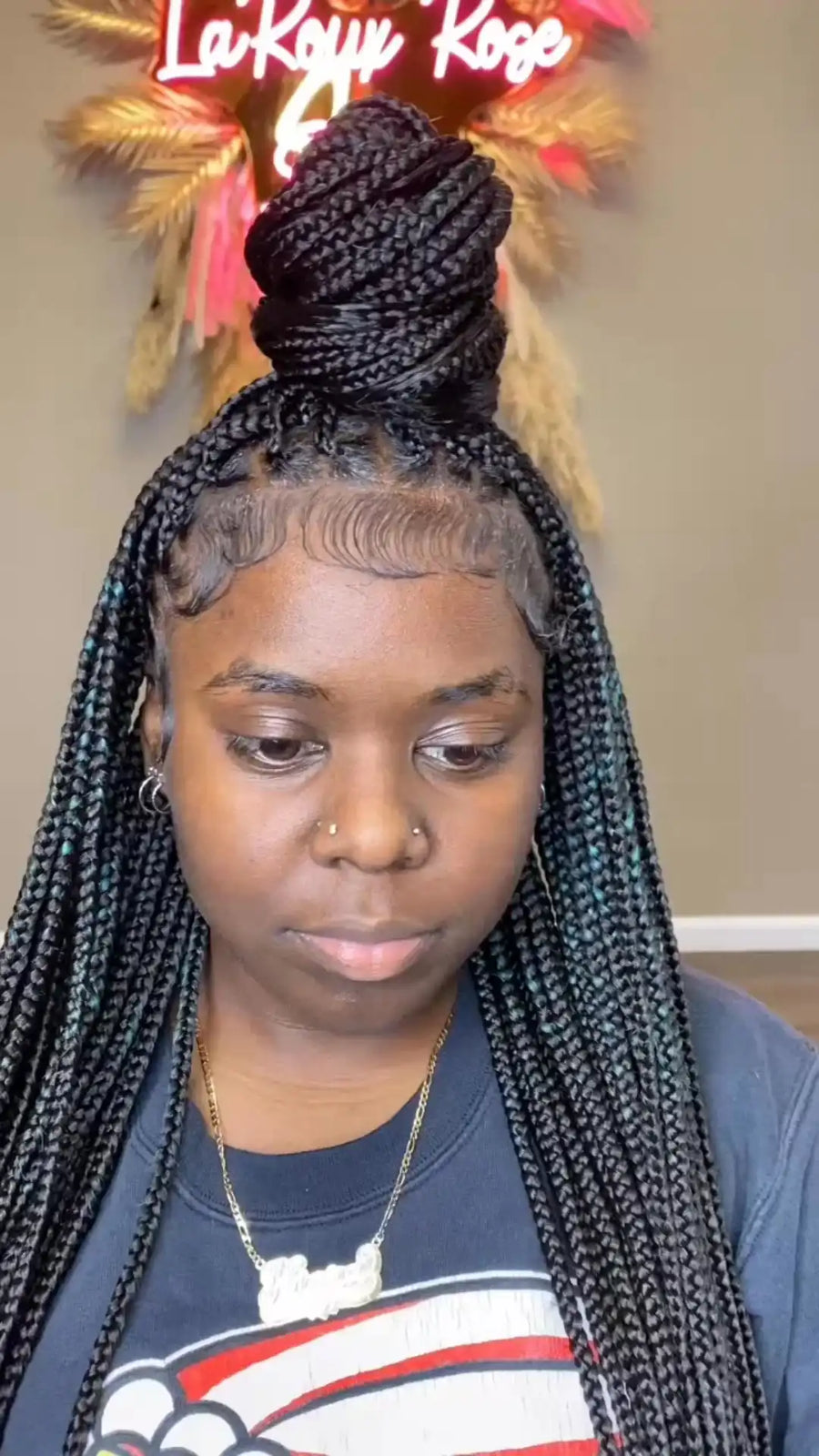 A person wearing black box braids styled in a high top knot with long side braids, shown from the chest up against an indoor wall with a decorative sign.