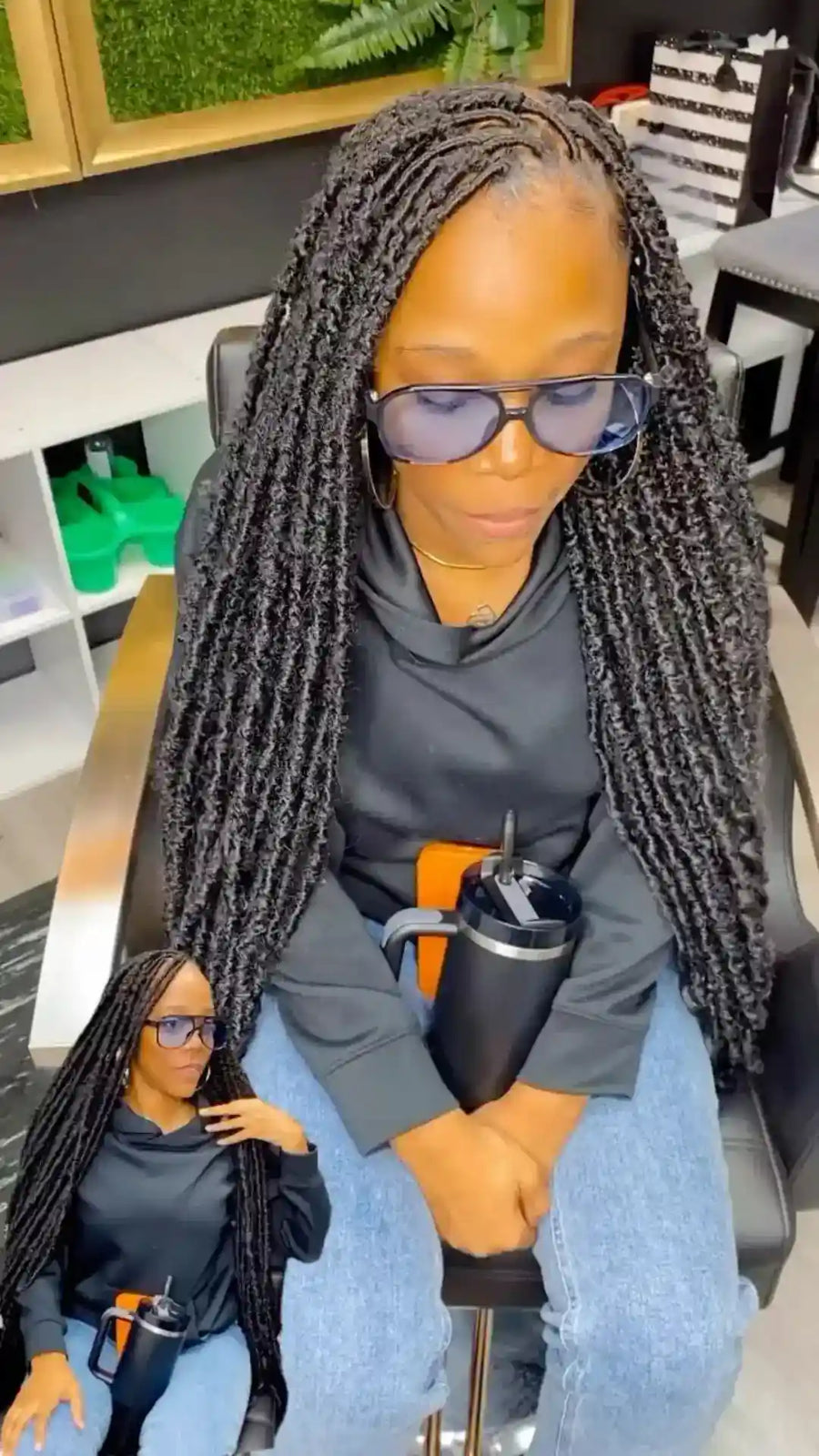 A woman with long black box braids sits in a salon chair wearing a dark gray hoodie and blue jeans, holding a black tumbler. A smaller inset image in the lower-left corner shows a doll or mannequin dressed to match her outfit and hairstyle.