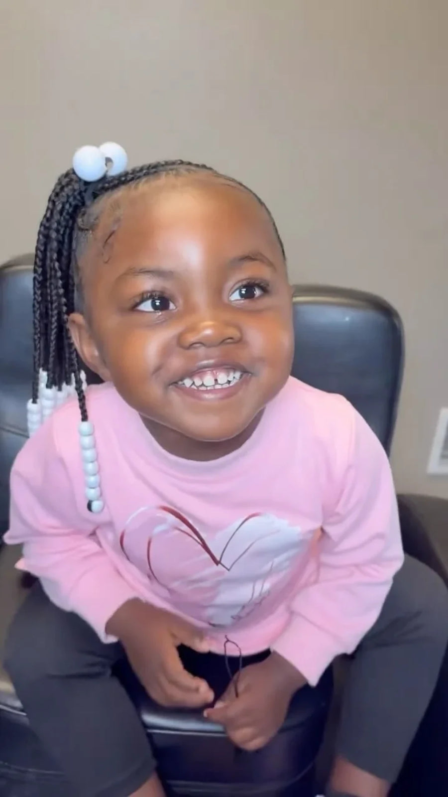 A young child with braided hair and white beads sits in a black chair, wearing a pink long-sleeve shirt with a white heart graphic and dark gray pants.