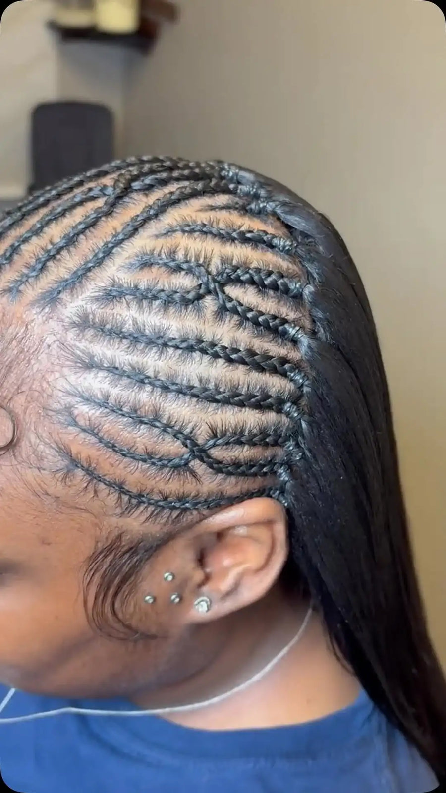 A close-up side view of a person with completed cornrow braids in dark brown or black hair, wearing a blue shirt and ear piercings, set against an indoor background.