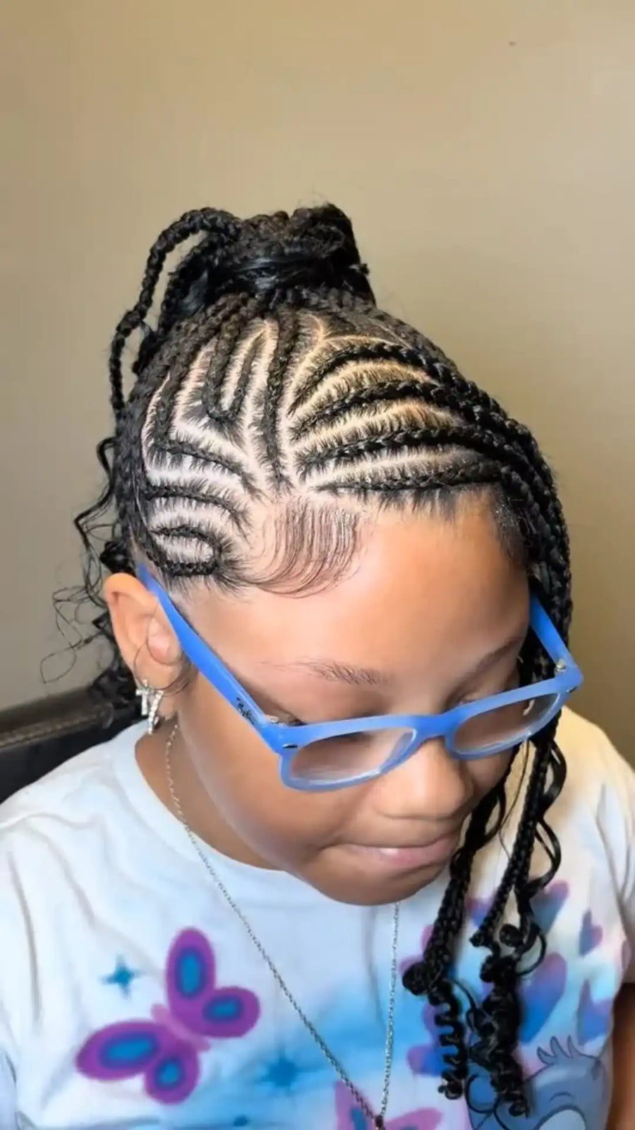 A close-up of a person with intricate cornrow braids styled in a high bun and loose ends, wearing blue-framed glasses and a white graphic t-shirt.