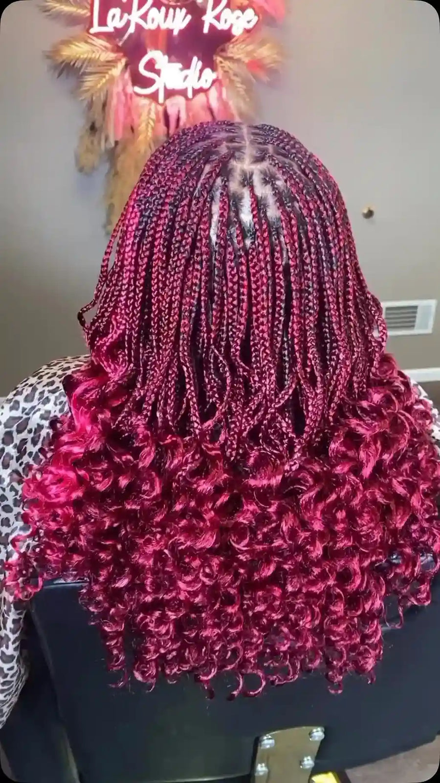 A person shown from the back wearing a long, curly wig in a deep magenta-pink color. The salon wall behind features a neon sign reading 'LaKouy Rose Studio'.