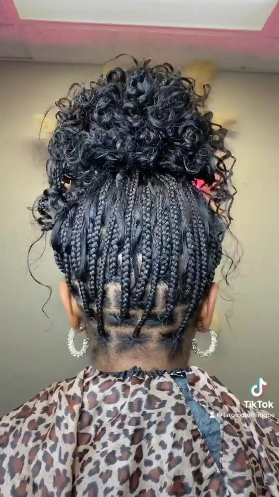 Back view of a person with a large black curly hair updo and braided cornrow extensions, wearing a leopard print top and silver hoop earrings.