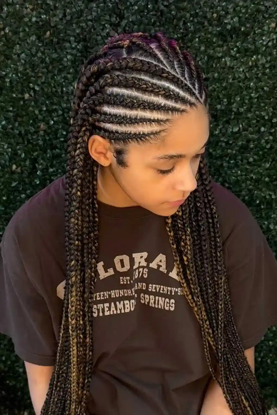 Person with braided hair wearing a dark t-shirt against a green leafy background