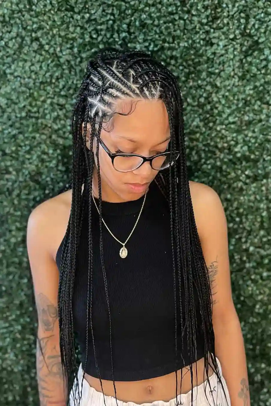 Person with braided hair wearing glasses and a black top against a green textured wall.