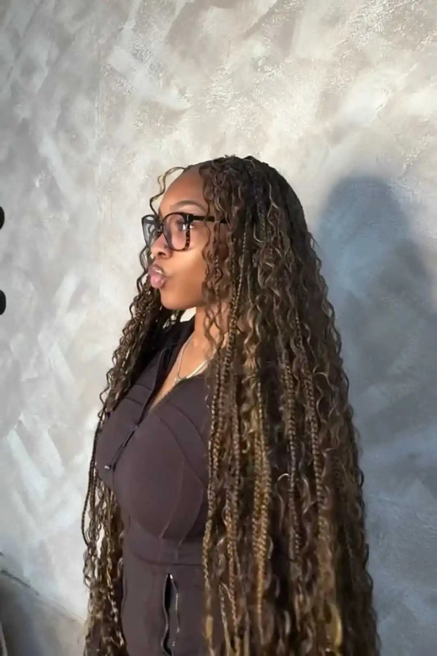 Woman with long braided hair wearing glasses against a textured wall.