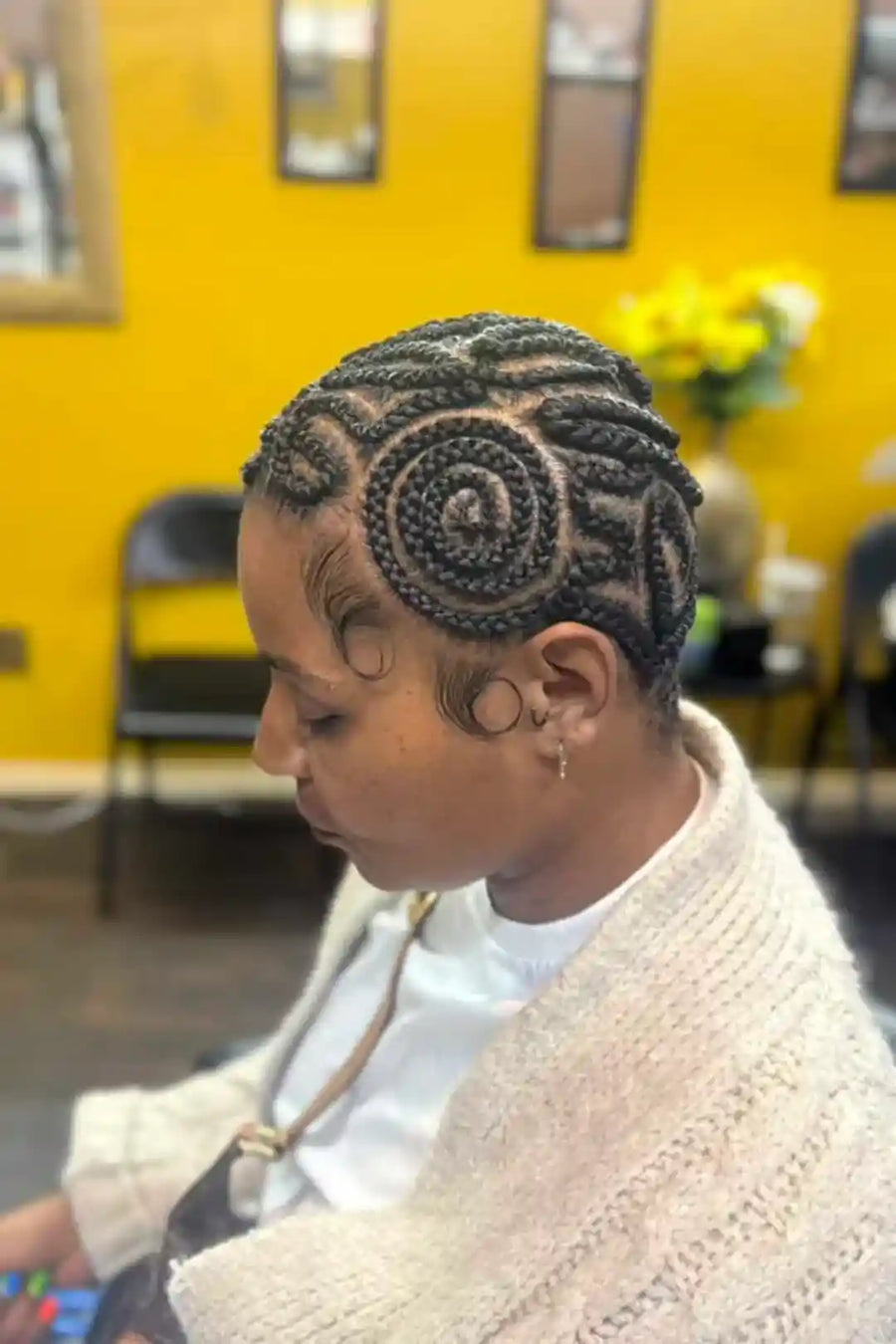 Person with intricate braided hairstyle in a room with yellow walls and chairs.