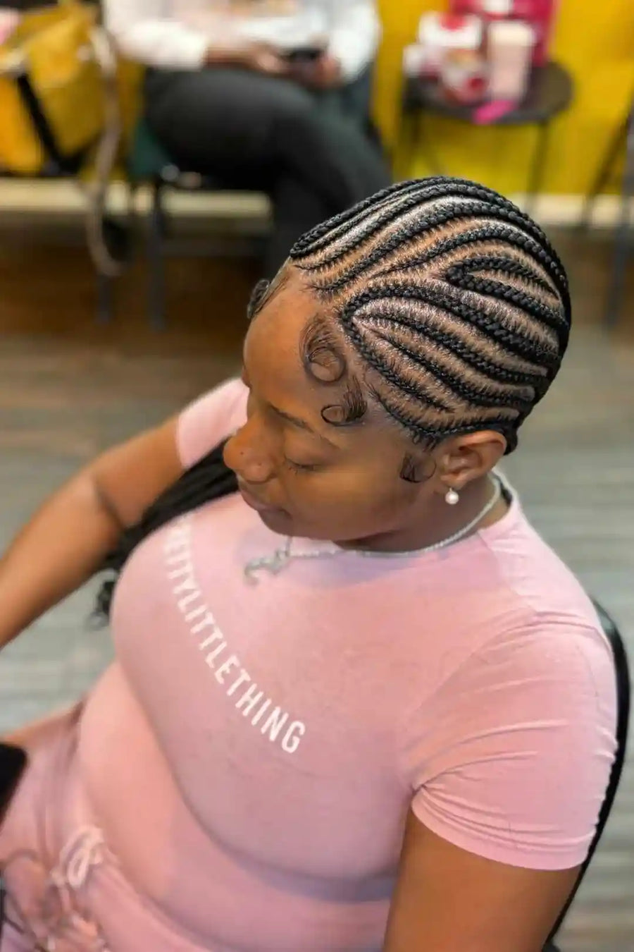 Woman with braided hair wearing a pink shirt with text, sitting in a casual setting.