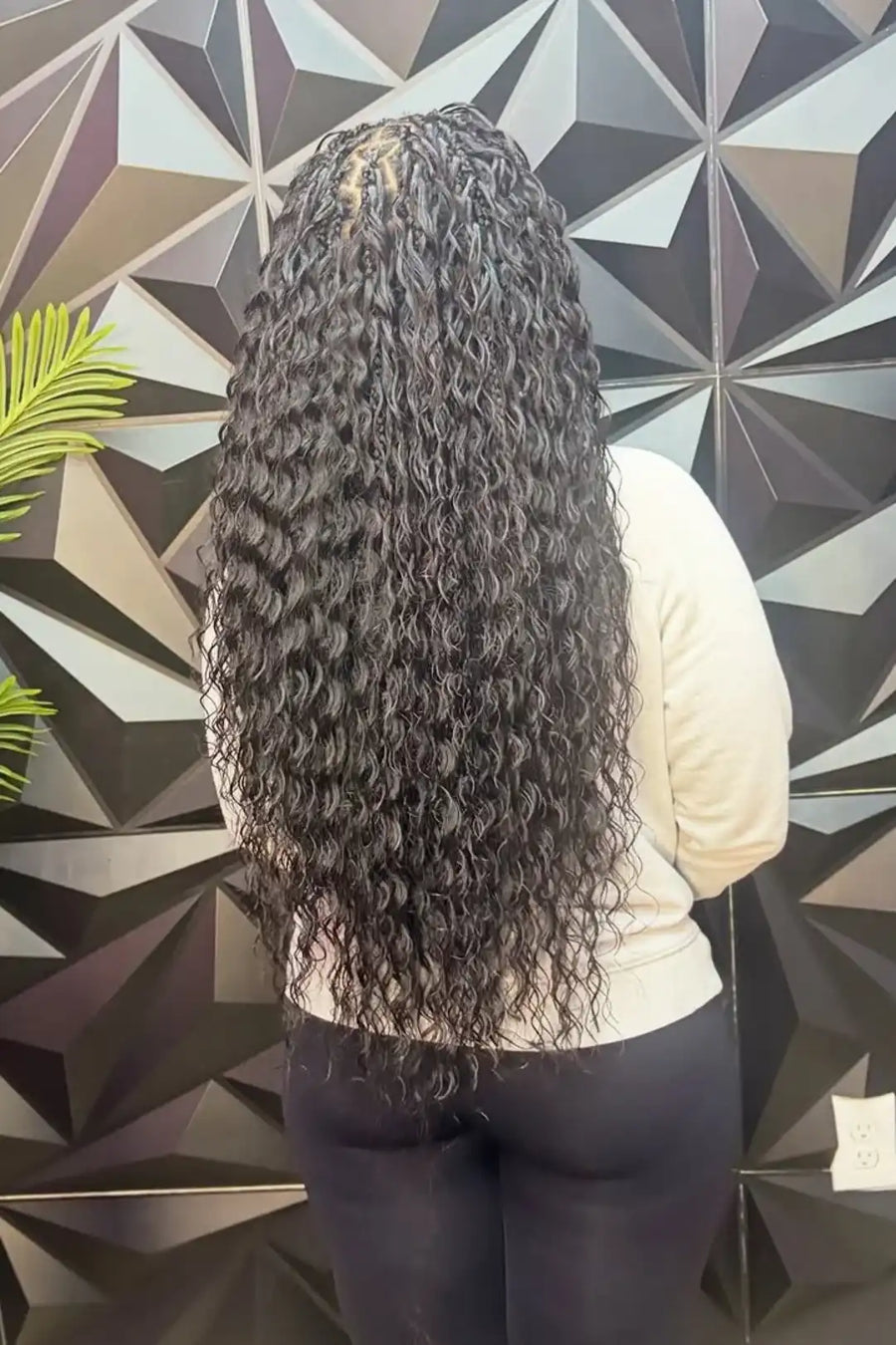 Person with long, curly hair standing against a geometric-patterned wall.