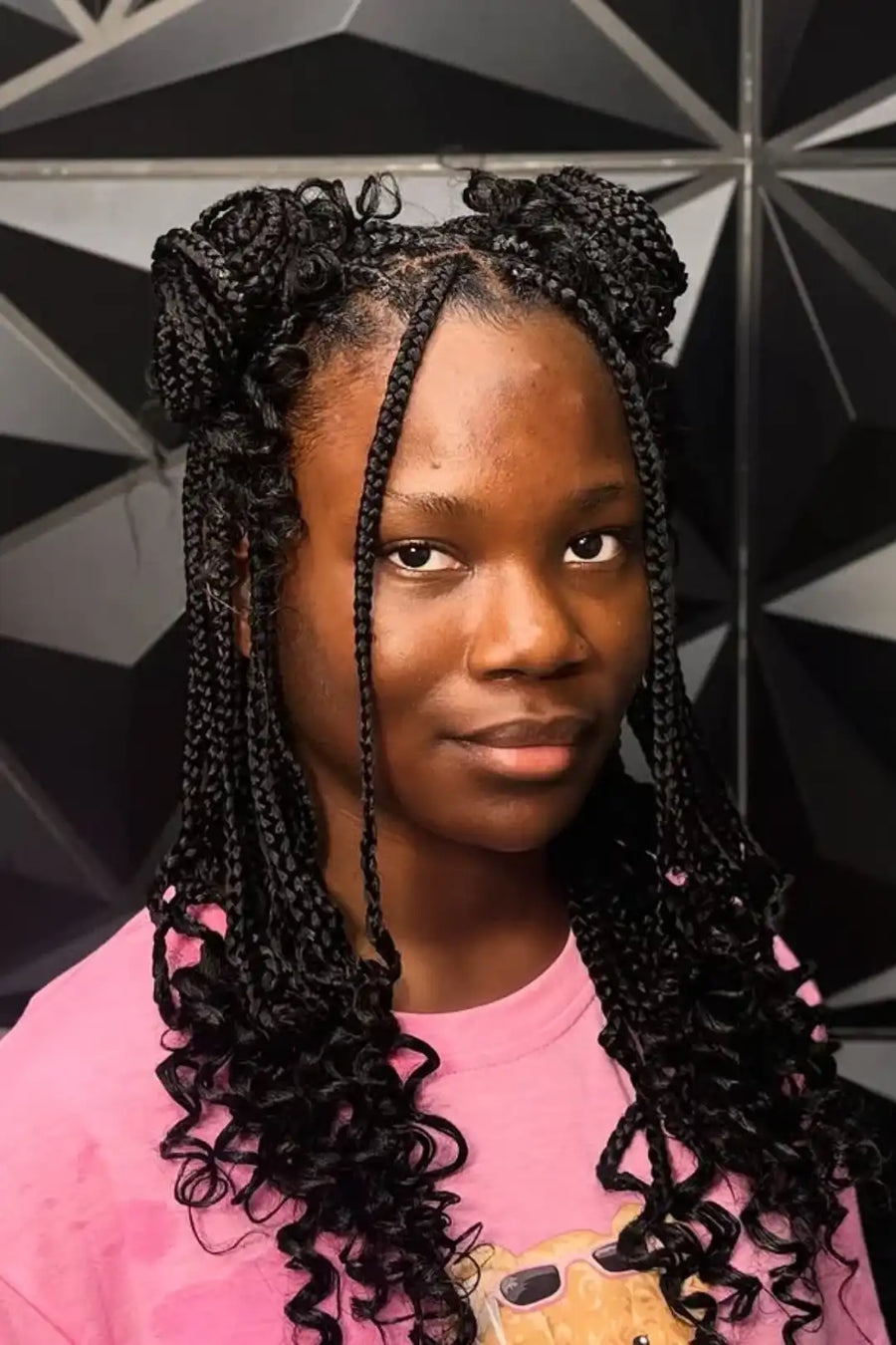 Person with braided hair wearing a pink shirt with a bear design against a geometric patterned wall.