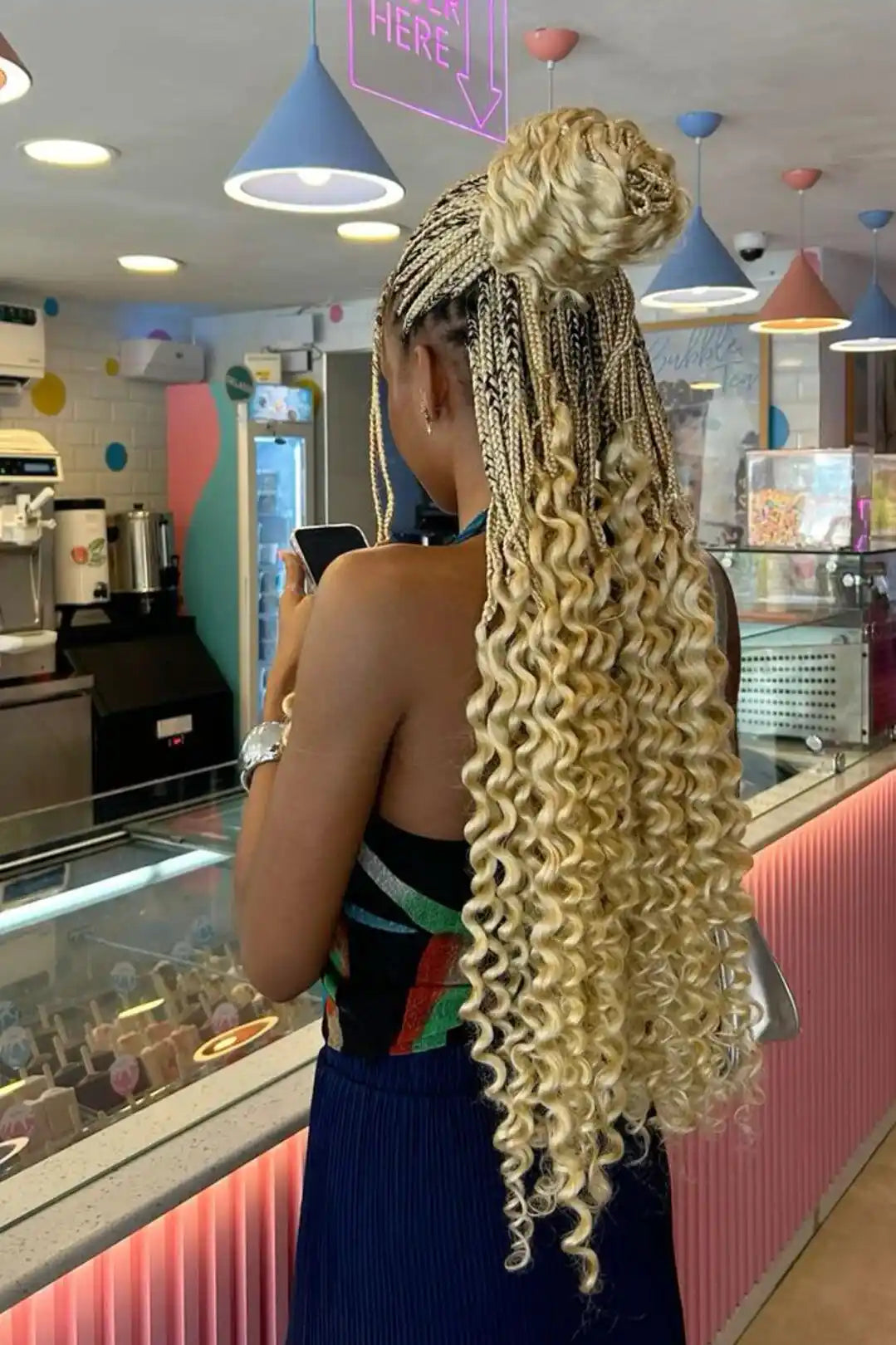 A woman standing in a colorful cafe or bakery, shown from the back, wearing a long blonde micro braided extension with a high bun top. The product is the synthetic braiding hair, which has a golden blonde color with darker brown and black accents.