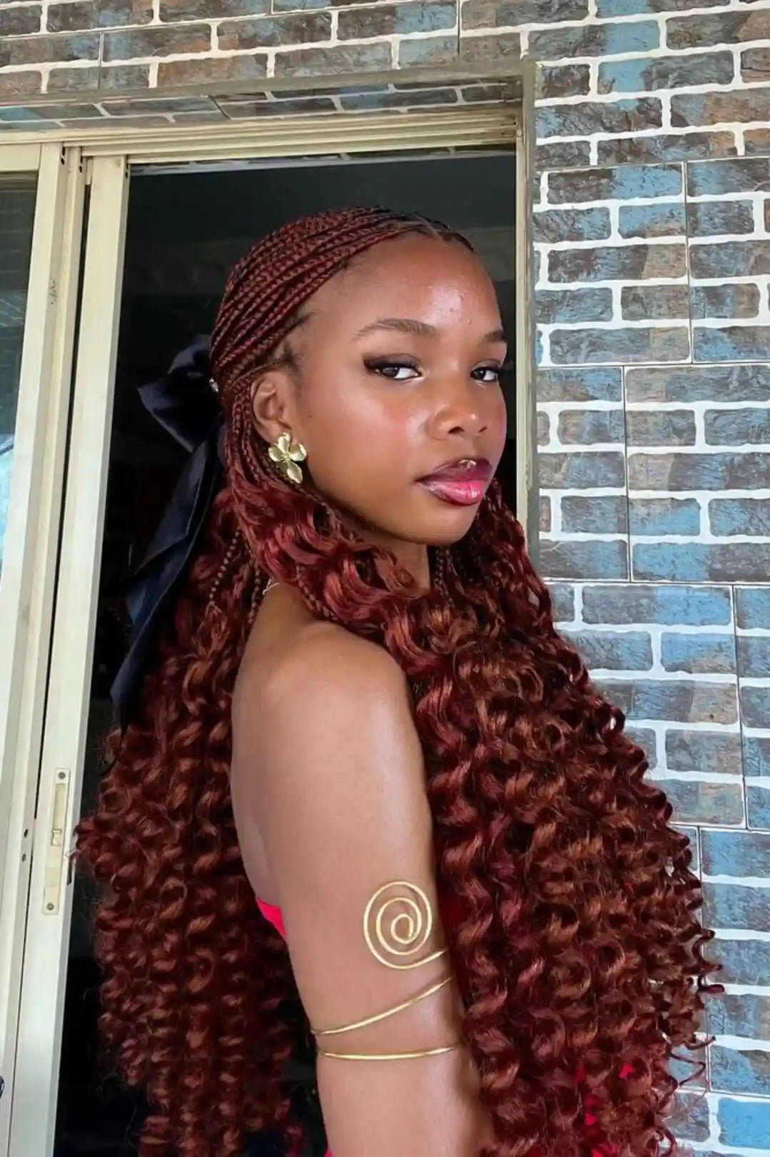 A woman with long, voluminous, deep auburn curly hair (or a wig) posing in front of a blue brick wall and doorway. She is wearing gold jewelry and a black hair bow.
