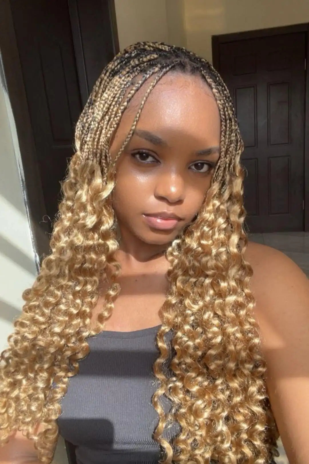 A woman with long, tight, and loose box braids in a blonde-to-brown gradient, wearing a dark gray ribbed tank top. She is taking a mirror selfie in an indoor setting with dark wooden doors in the background.