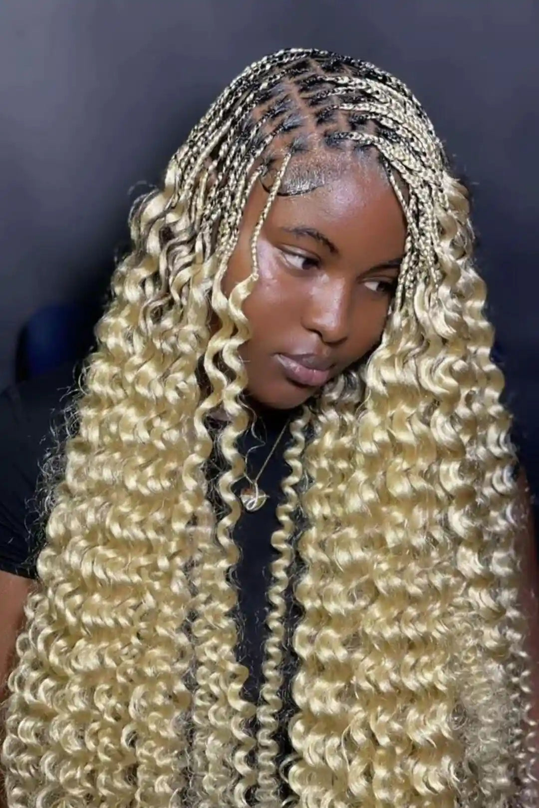 A close-up of a person wearing long, voluminous curly hair extensions in a light blonde shade, styled with braided cornrow tops and small gold chain accents against a dark background.