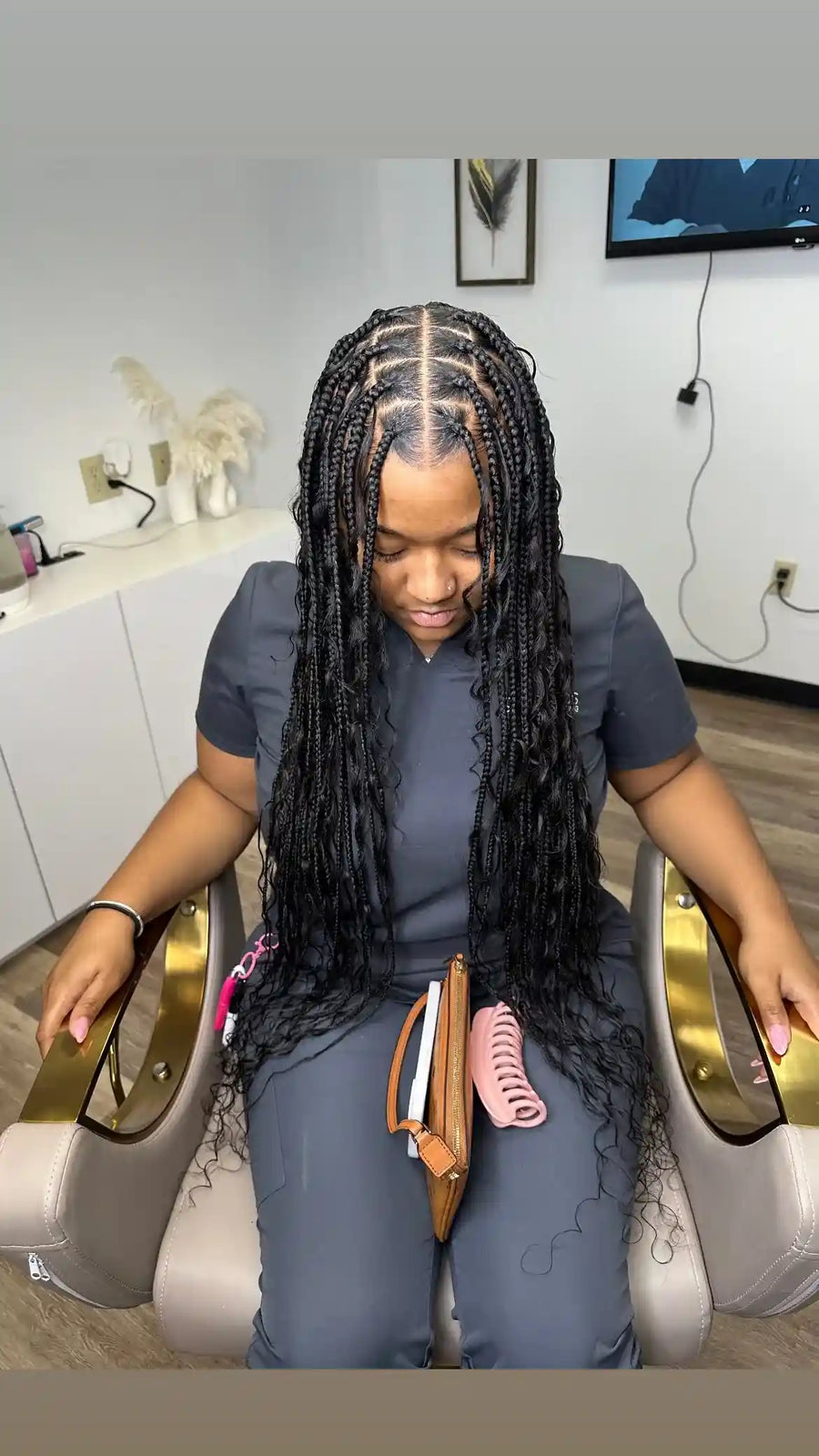 A woman with long, black, curly cornrow braids sits in a salon chair, wearing a dark gray scrub top and pants. She has a small brown leather pouch with pink hair clips resting on her lap.