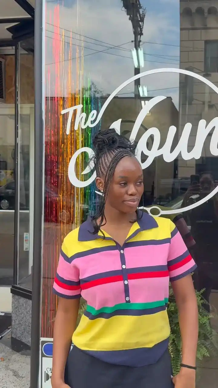 A woman wearing a colorful short-sleeve striped polo shirt with pink, yellow, green, and navy horizontal stripes, standing in front of a storefront window with 'The Coun' visible on it.
