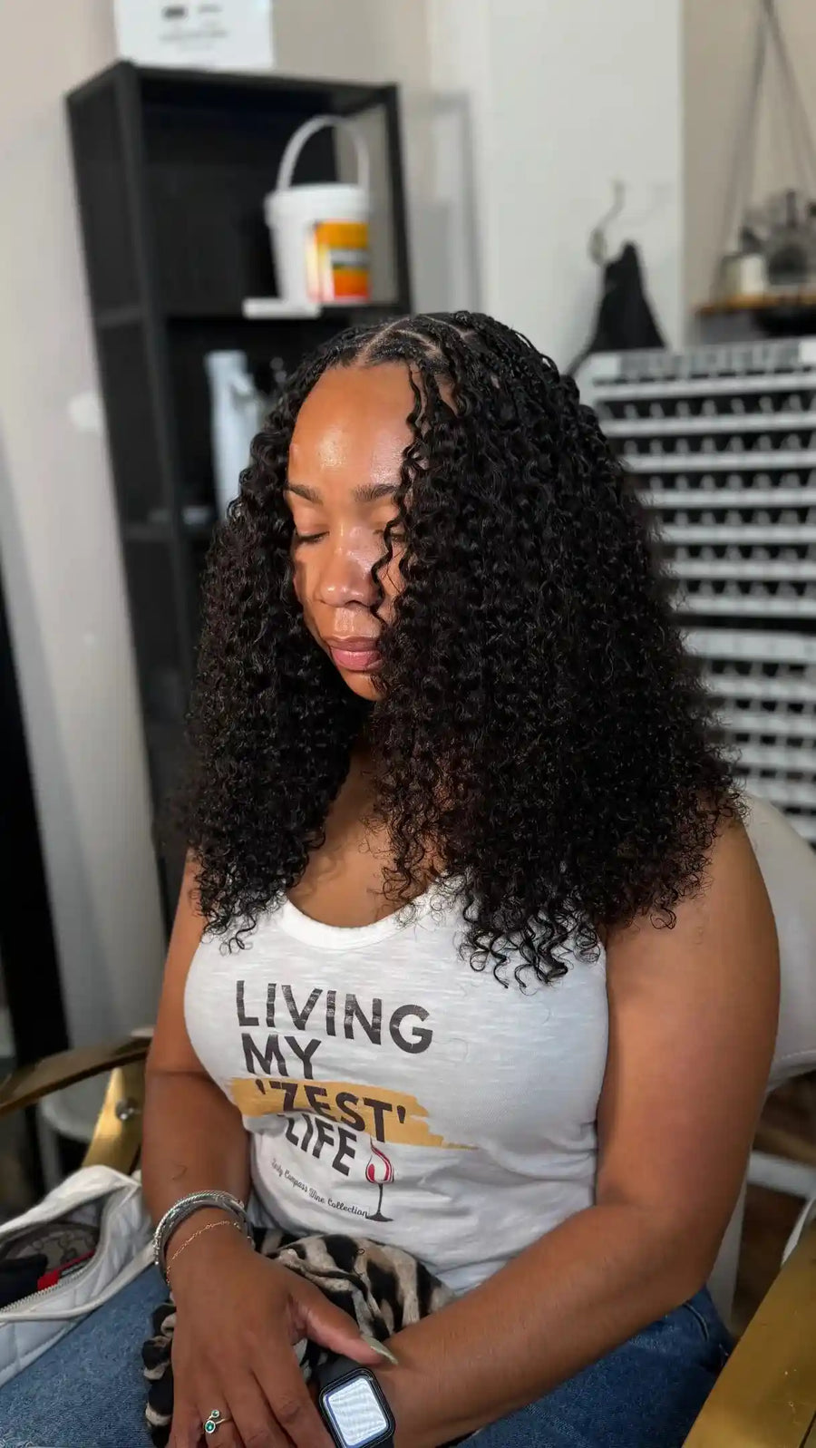 A woman with long, curly black hair sits indoors wearing a white graphic tank top that reads 'LIVING MY BEST LIFE.' The product appears to be the tank top rather than the hair, since the shirt is centered and prominently shown with printed text.