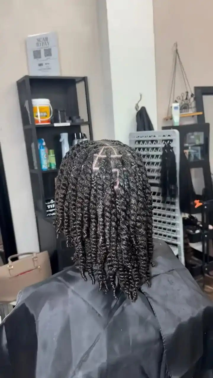 A person seated in a salon or hair studio, viewed from behind, wearing a black salon cape. The visible product appears to be a black twisted or braided hair extension/wig with tight curls.