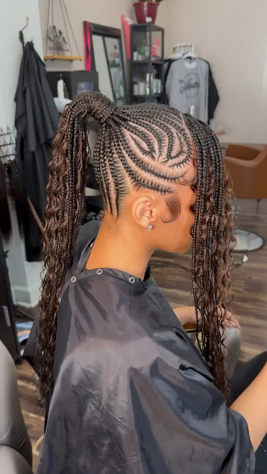 A person seated in a salon chair wearing a black salon cape, shown from the side/back with long dark curly hair styled in a high ponytail and intricate braided cornrow patterns across the scalp. The salon interior is visible in the background.