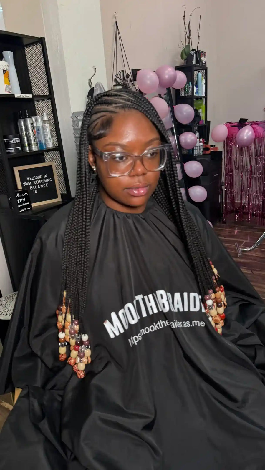 A person with long black braids sits wearing a black salon cape printed with the white text 'MOUTHBAID' and a website, in what appears to be a salon or studio setting with shelves, bottles, and pink balloons in the background.