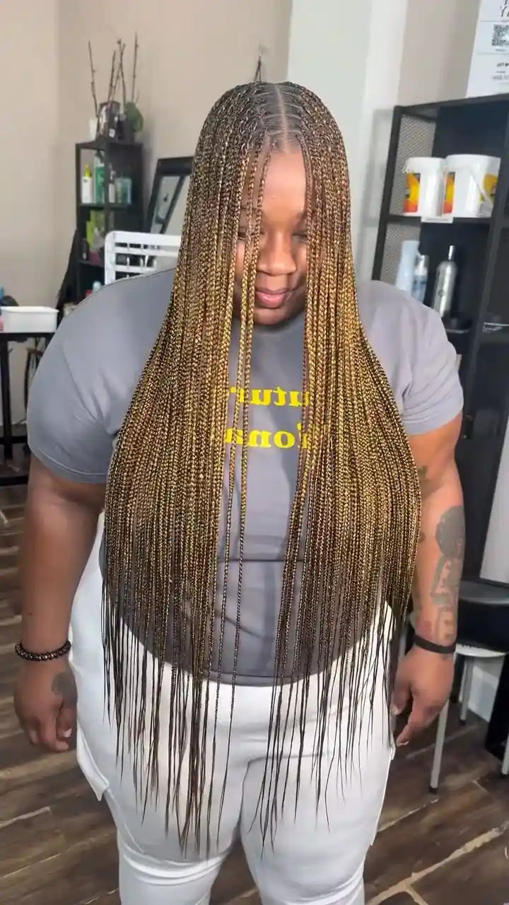 A person standing indoors wearing a gray T-shirt with yellow text and white pants, featuring very long, straight braided hair in a light brown to dark brown color.