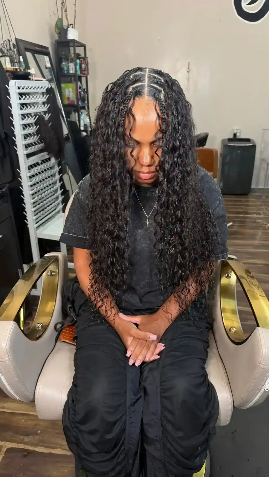 A person sitting in a salon chair wearing a long, black, curly wig with a center part down the middle. The wig fills most of the frame, and the background shows a salon interior with shelves and equipment.
