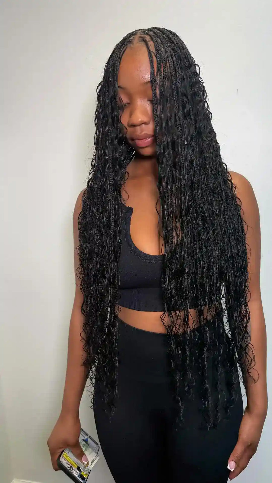 A woman with long, black, curly micro braids or a wig wearing a black sports bra and leggings, holding a phone in her hand against a plain white wall.