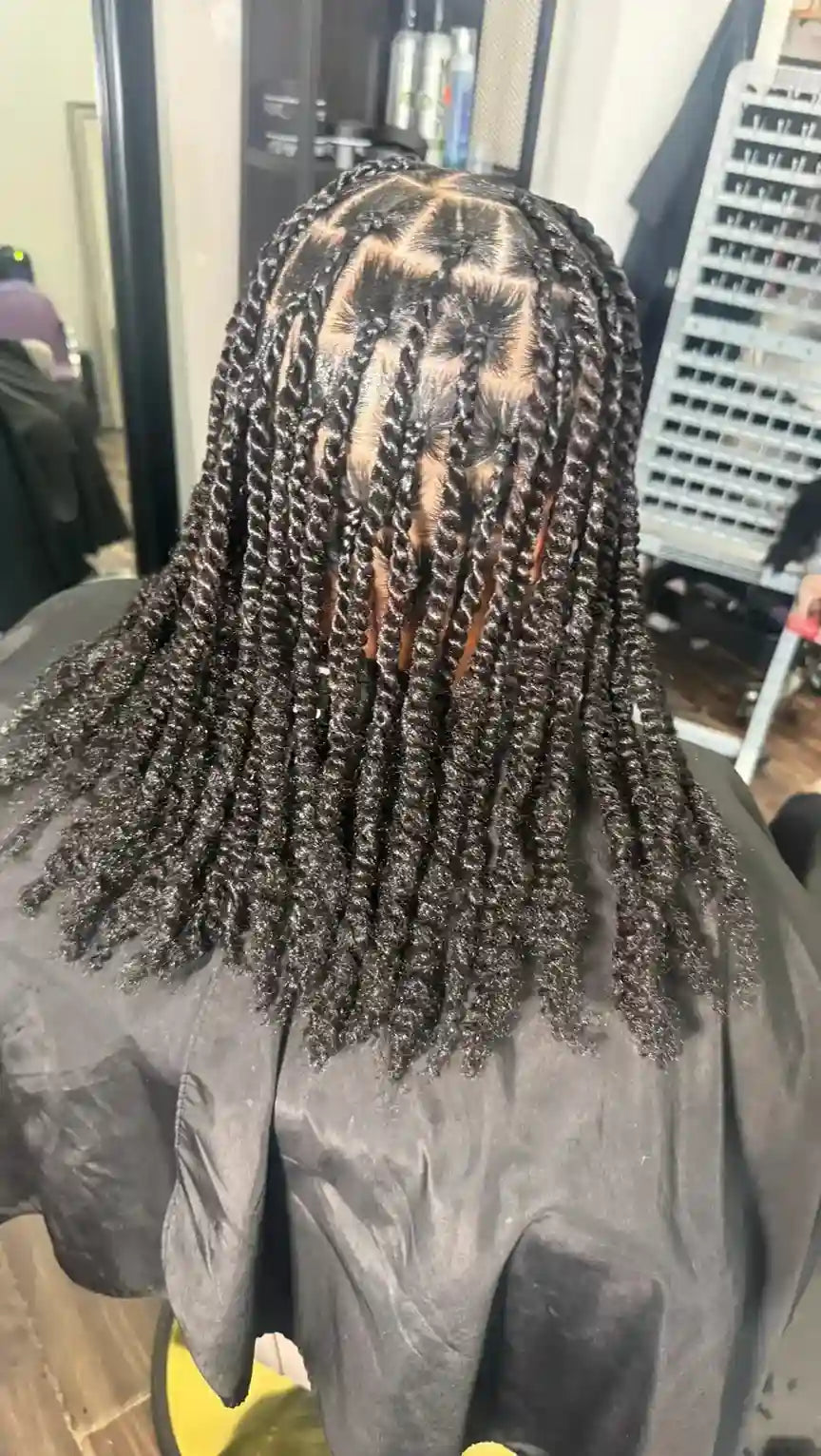 Back view of a person with long, tightly curled black braids or box braids, seated in a salon with a black salon cape visible.