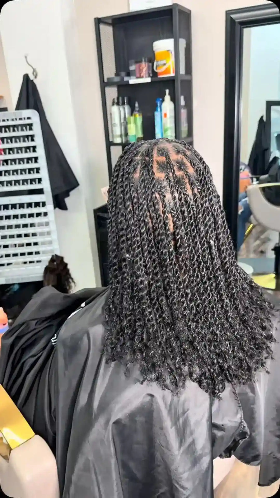 A person seated in a salon chair viewed from behind, wearing a black salon cape and showing long, tightly curled black hair. A salon interior with shelves, bottles, a mirror, and chair is visible in the background.