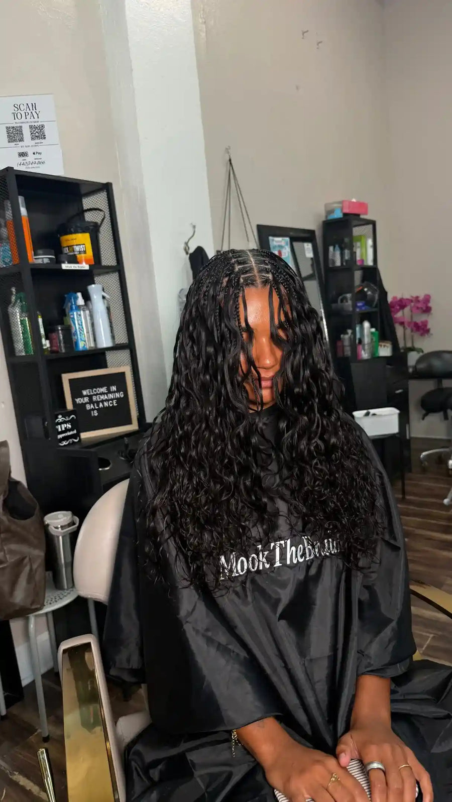 A person sitting in a salon chair wearing a black hair styling cape with the text 'Mook The Beauty' printed on the front. The person has long, dark, curly hair and is photographed inside a salon with shelves and mirrors in the background.