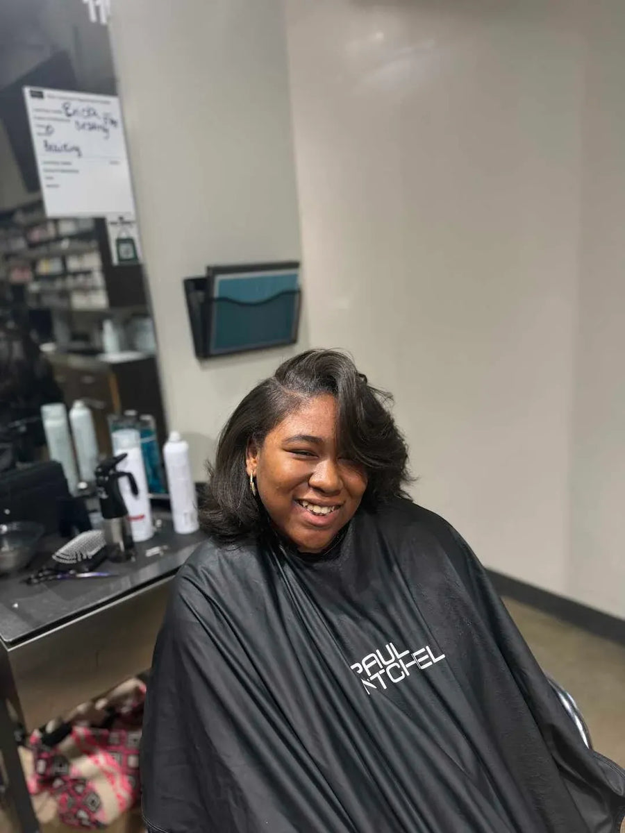 Person sitting in a salon chair wearing a black 'Paul Mitchell' cape.