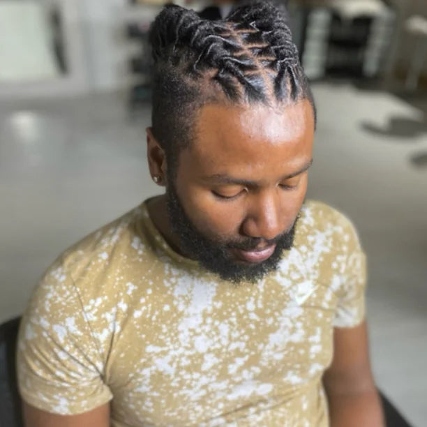Man with braided hair wearing a beige t-shirt with white patterns.