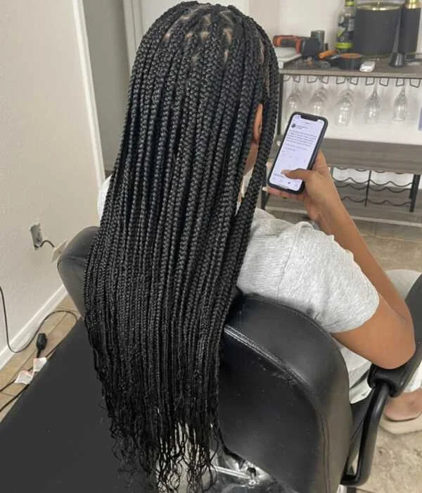 Person with long braided hair sitting in a salon chair using a phone.