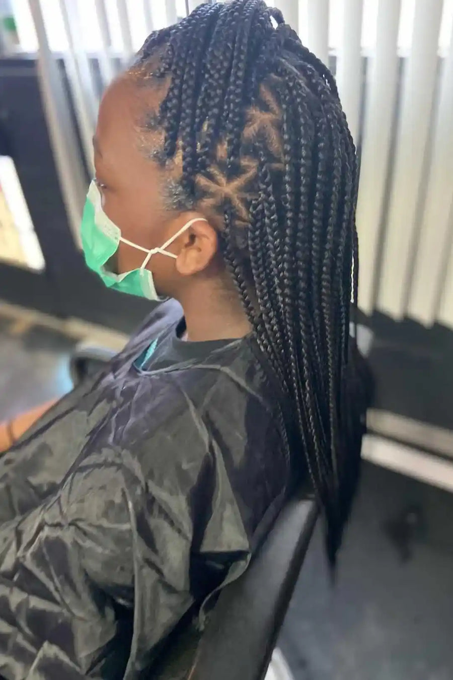 Person with braided hair wearing a face mask and sitting in a chair.