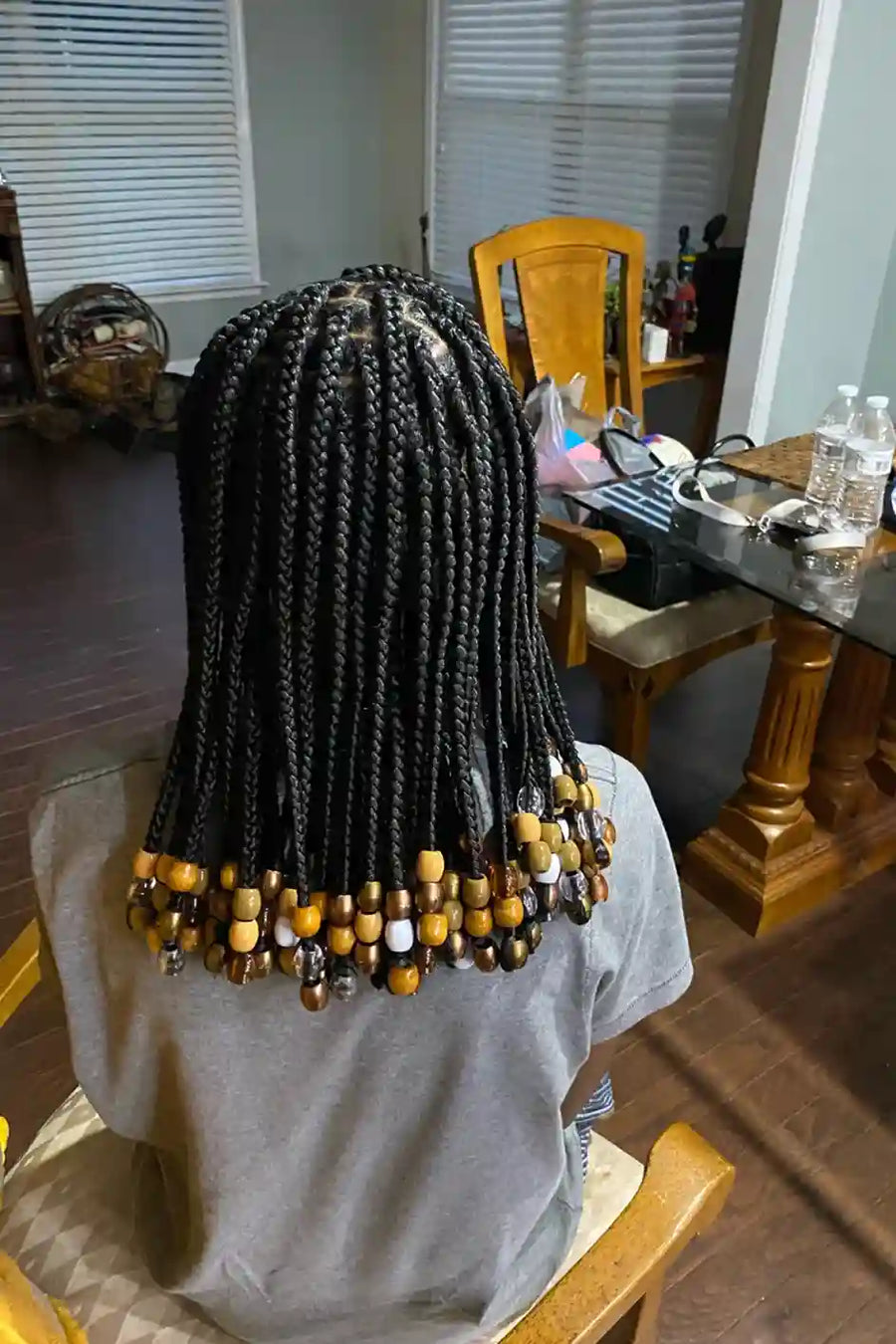 Person with braided hair and beads in a room with wooden furniture and a window.