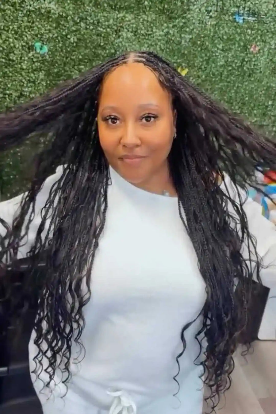 Woman with long, styled hair wearing a white top against a green textured wall.