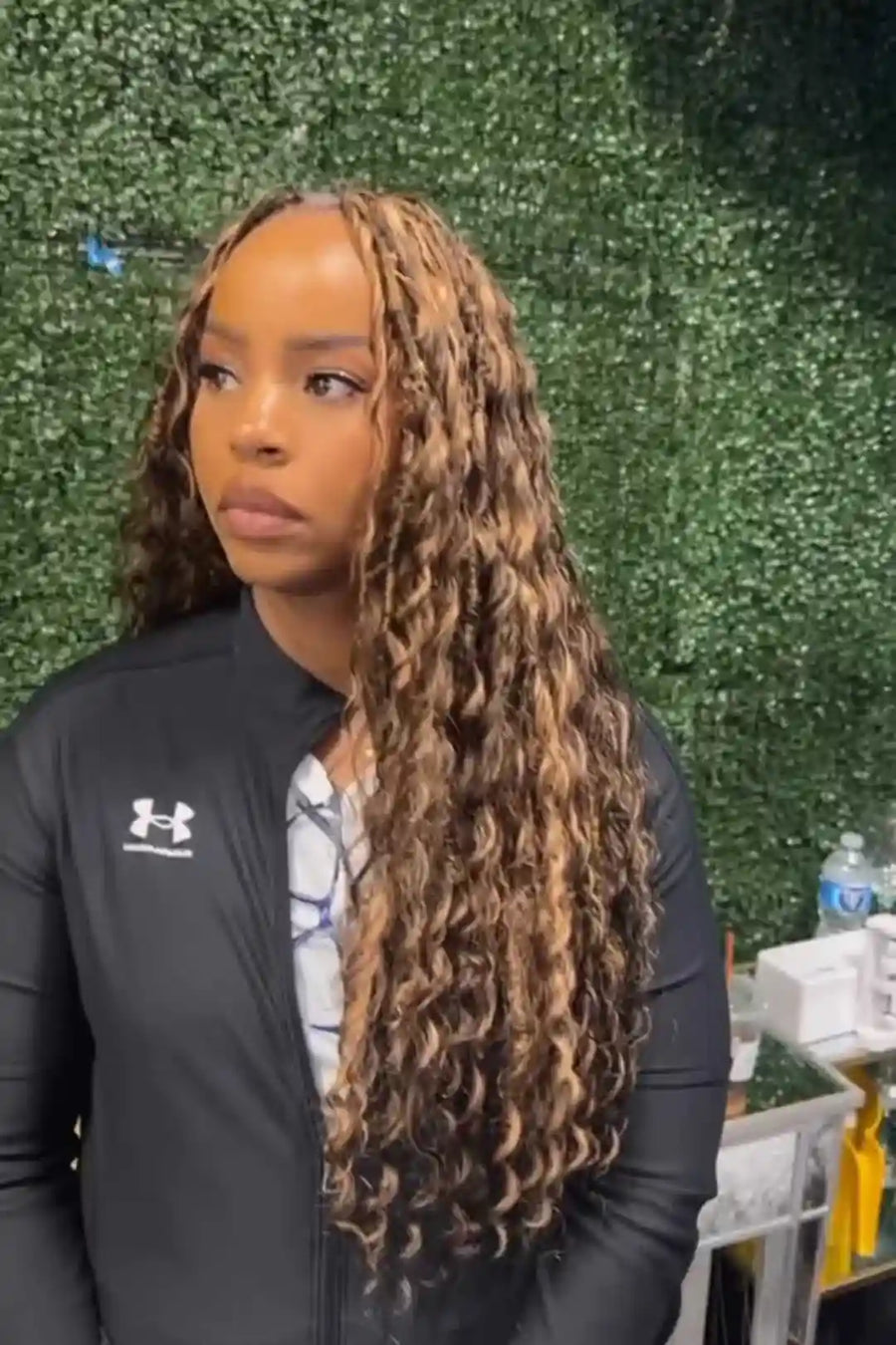 Person wearing a black Under Armour jacket with long braided hair against a green leafy background.